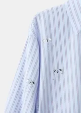 Camisa de popelina celeste a rayas, de corte oversize, con cuello camisero, mangas largas y aplicaciones de pedrería y lentejuelas brillantes distribuidas por toda la prenda.