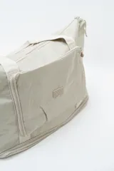 Bolso de viaje tipo duffle, color beige, confeccionado en material sintético ligero. Cuenta con doble asa de mano, cierre principal superior, bolsillo frontal con cierre y compartimento inferior independiente con cierre.