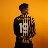 Camiseta de fútbol Puma de manga larga, conmemorativa del aniversario del Club Atlético Peñarol. Diseño de estilo retro que combina un sector con rayas verticales en amarillo y negro, y otro sector liso en negro, con cuello polo y detalles en dorado.