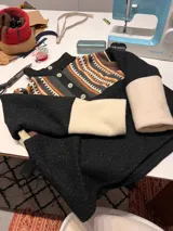 Buzo negro de cuerpo texturizado con mangas largas que combinan tejido negro y puños en color crudo. Presenta cuello alto con botonera frontal de cuatro botones y detalle estampado étnico multicolor en la parte superior.