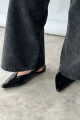 Zapato tipo flat destalonado de color negro con acabado brillante, punta fina y cierre con hebilla lateral.