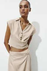 Camisa de lino con cuello solapa y escote en pico. Presenta un diseño sin mangas con detalle de pliegue cruzado en el delantero y cierre frontal con un botón.