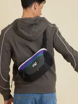 Bandolera deportiva Rusty con diseño de bloques de color en negro, violeta y verde. Cuenta con doble cierre, compartimentos de malla lateral y correa ajustable.