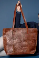 Bolso tipo tote confeccionado en cuero vacuno con textura de reptil, color marrón. Presenta costuras decorativas en los bordes, doble asa de hombro y un detalle de borla de flecos de cuero colgante.
