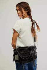 Cartera de cuero color negro con efecto gastado, de cuerpo blando, asas dobles de mano y correa cruzada desmontable. Cuenta con bolsillos exteriores con cierre, tiras colgantes decorativas y un dije metálico de la marca We The Free.