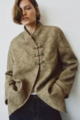 Blazer de corte recto confeccionado en tejido jacquard con diseño floral en tonos tostados. Presenta cuello mao, manga larga y cierre frontal mediante alamares decorativos. Incluye bolsillos de vivo en el delantero.