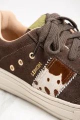 Zapatillas bajas de gamuza color marrón con cordones y suela de goma color crema. Presentan una franja lateral con estampado de vaca y el logo de la marca bordado en el lateral.