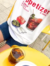 Buzo de manga larga color blanco, corte oversize con hombros caídos. Estampado frontal con la palabra "appetizer" en rojo y una ilustración de un cóctel con aceitunas y rodaja de naranja, acompañado de pequeños cuencos con snacks.