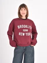 Buzo de cuello redondo en color bordó con estampado frontal en blanco que incluye el texto Brooklyn 11208 New York. Presenta un diseño de corte holgado con hombros caídos y puños acanalados.