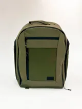 Mochila de color verde oliva con paneles en tonos de verde contrastantes y detalles en negro. Presenta un bolsillo frontal con cierre y un pequeño parche rectangular con texto en la parte inferior.