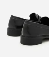 Mocasín Satinato de mujer confeccionado en cuero negro con acabado liso, puntera redondeada, costuras visibles, tira decorativa en la pala y suela con taco bajo.