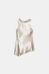 Top satinado color beige con cuello fluido, manga sisa y aplicaciones de brillos. Presenta falsos botones forrados en el lateral, bajo asimétrico con abertura y cierre de cremallera oculta.