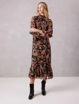 Vestido midi de gasa con estampado de arabescos en tonos cálidos sobre fondo negro. Presenta cuello alto con detalle de punto smock, mangas largas con puños elásticos y volados, y falda con tajo lateral y ruedo con volado. Incluye forro interno y cierre posterior.