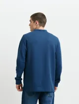 Remera de manga larga con cuello polo, confeccionada en tejido de punto canalé francés. Presenta abotonadura frontal con botones de madera y detalles a contratono en la cartera y el interior del cuello.
