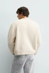 Cárdigan de punto color beige con cuello en pico, manga larga y cierre frontal de botones. Presenta un pequeño bordado a contraste en el pecho y acabados en rib en puños y bajo.