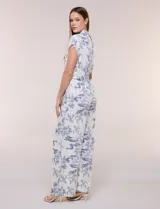 Enterito largo color blanco con estampa ilustrada en azul estilo Toile de Jouy, escote en V cruzado, mangas cortas caídas, bolsillos laterales, pantalón amplio wide leg y cinturón incorporado con hebilla metálica.