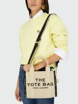 Bolso tipo tote de tamaño pequeño, confeccionado en tejido jacquard color beige con detalles en negro. Presenta el nombre del modelo estampado en el frente, doble asa superior y correa de hombro ajustable y extraíble con diseño de cinta.