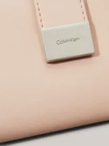 Billetera mediana tipo bifold de piel sintética estructurada color rosado pálido, con cierre mediante solapa y broche metálico a presión. Presenta una placa metálica con el logo de Calvin Klein en el frente.