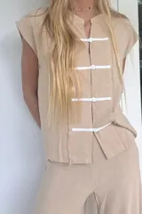 Blusa sin mangas color beige, con cuello mao y cierre frontal con botones de nácar y tiras de tela blanca. Presenta un acabado deshilachado en los bordes.
