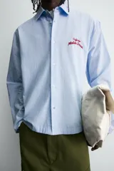 Camisa de algodón de corte regular, color celeste con finas rayas blancas verticales. Presenta un bordado en color rojo en el pecho con la frase "Café du Matin Doux".