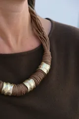 Collar de diseño artesanal compuesto por múltiples hilos de cuero color chocolate, adornado con piezas cilíndricas de metal dorado con textura irregular.