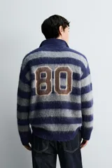 Polo de punto cepillado con diseño de rayas horizontales en azul marino y gris. Presenta cuello solapa con abertura frontal, manga larga y texto bordado en el pecho. Acabados en rib en puños y ruedo.