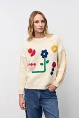 Sweater de punto color crudo con diseño de flores y formas geométricas bordadas en colores vibrantes. Presenta cuello redondo, mangas largas con detalles de líneas laterales en contraste y corte holgado.