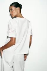 Camiseta de algodón de corte regular, color blanco, con cuello redondo y manga corta. Presenta detalles de bordados florales y ornamentales en color rosado en el ruedo y en las mangas.