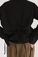 Camisa negra de lino con cuello camisero, manga larga y cierre frontal con botones de nácar. Presenta un diseño con cordones laterales que permiten fruncir la prenda para un ajuste personalizado.