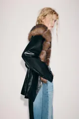 Trench corto Zara de efecto piel color negro, con cuello de solapa y aplicación de pelo sintético a contraste en color marrón. Cuenta con manga larga con trabillas en los puños, bolsillos delanteros y cinturón ajustable del mismo tejido.