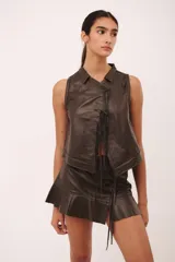 Falda corta de cuero color marrón oscuro, con ruedo con volado y detalles de cordones ajustables en el frente y laterales.