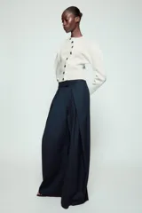 Pantalón de tiro medio de corte wide leg, confeccionado en mezcla de lyocell con patrón de rayas diplomáticas. Presenta bolsillos laterales ocultos, pliegues delanteros marcados y cierre frontal con cremallera, botón y ganchos metálicos.