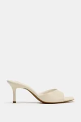 Sandalia de tacón tipo mule color beige, con tira ancha delantera, tacón fino alto y punta redondeada.