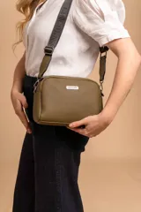 Bandolera de diseño compacto con textura granulada, color verde oliva. Cuenta con una correa ajustable de tela negra con el logo de la marca en relieve y placa metálica frontal con el nombre de la marca.