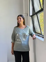 Remera de manga larga color verde, con cuello redondo y diseño holgado. Presenta un detalle frontal con parches de lentejuelas plateadas y estampado tipográfico.