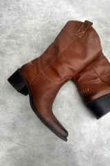 Bota de cuero color marrón con caña media, bordado a contratono, tacón bajo, punta alargada redondeada y tiradores laterales.