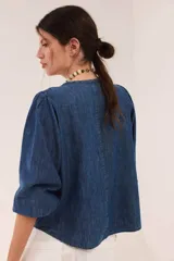Blusa de jean fino de algodón azul, con escote a la base, mangas abullonadas y puños elásticos. Cuenta con pechera decorada con cintas superpuestas de efecto rústico y desmechado.