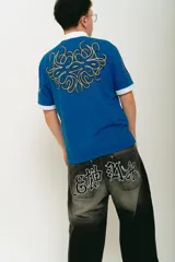 Pantalón de jean negro con lavado localizado (efecto tie-dye o acid wash), corte baggy y amplio. Presenta apliques bordados en la parte trasera de los bolsillos y herrajes metálicos cromados.