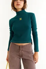 Top de manga larga con cuello alto, confeccionado en tejido de lycra y algodón. Presenta costuras a contratono, bordado central en el pecho, aberturas laterales en el ruedo y diseño con dedal en las mangas.