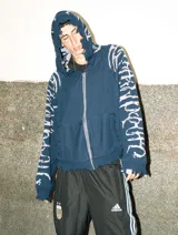 Hoodie con cierre frontal, tejido en algodón con diseño reversible. Presenta detalles de roturas intencionales en puños, ruedo y costuras, con estampado gráfico en mangas y capucha.