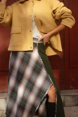 Falda midi de diseño asimétrico que combina un panel de tela escocesa en tonos verdes, blancos y marrones con un panel lateral de paño verde oscuro. Presenta un tajo lateral pronunciado y tiro a la cintura.