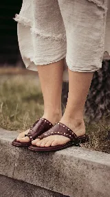 Sandalias chatas de cuero color bordó, con diseño de tira ancha que envuelve el empeine y sujeta el dedo gordo, decoradas con tachas metálicas doradas.