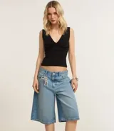 Bermuda tipo jorts de tiro medio, confeccionada en denim de algodón color azul claro. Presenta un corte holgado que llega por debajo de la rodilla, con trabillas en la cintura, bolsillos laterales y traseros, y un detalle de cordón decorativo en la parte frontal.