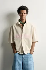 Sobrecamisa boxy fit de algodón color beige, de manga corta, con cuello solapa, cierre frontal de botones y aplicación de texto en parches rosas a contraste en el delantero.