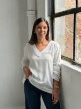 Blusa blanca de corte holgado con escote en V, mangas largas y costuras visibles en el cuello y el ruedo.