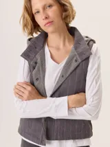 Chaleco gris con cuello alto y capucha, cierre frontal con botones a presión. La parte delantera es de lana gris lisa y la parte interior del cuello y el forro son de tela con patrón de espiga en blanco y negro.