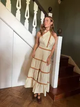 Vestido midi de lino color beige, con diseño de cuello halter cruzado y detalles de volados en tono marrón en el cuello, cintura y los niveles de la falda.