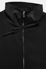 Chaleco fluido color marrón con cuello subido, sin mangas. Presenta cierre frontal de cremallera metálica y detalles de lazadas ajustables en el cuello y la cintura.