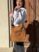 Cartera tipo tote bag de cuero color camel, con detalles de tachas metálicas en el bolsillo frontal y tiras largas para el hombro.