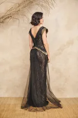 Vestido largo de encaje negro con capas de tul, escote en V cruzado, mangas cortas con detalles metalizados en dorado y falda de corte sirena con cola y sobrefalda drapeada.